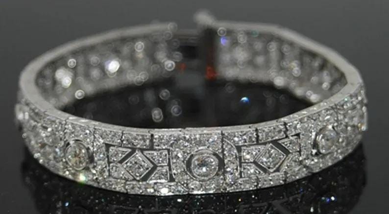 May include: A silver-toned metal bracelet featuring a repeating pattern of circular and geometric links. The entire surface is encrusted with numerous small, clear, sparkling stones set in a dense, intricate arrangement across the band.