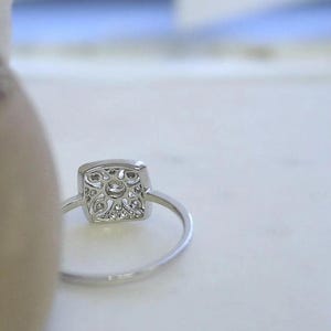K&ouml;nnte beinhalten: Ein silberner Ring mit quadratischer, verzierter Fassung. Die Fassung ist mit kleinen, klaren Edelsteinen um einen gr&ouml;&szlig;eren zentralen Stein besetzt. Der Ring ist schmal und poliert. Der Ring ist vor einem weichen, verschwommenen Hintergrund platziert.