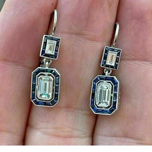 May include: A pair of silver drop earrings with rectangular and emerald-cut clear stones. The earrings have a dark blue border around the stones. The earrings are hanging from a silver hook.