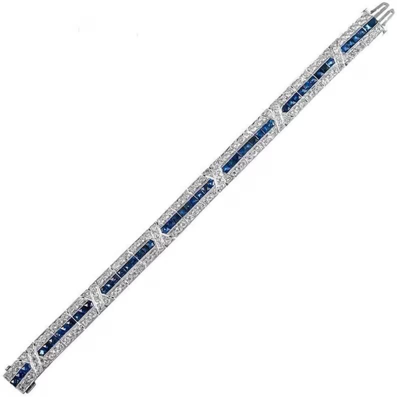 May include: A silver-toned metal bracelet featuring a central row of square-cut blue sapphires flanked by two rows of small, round, clear white stones. The design includes repeating X-shaped metal links set with clear stones between the sapphire segments.