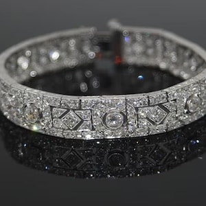 May include: A silver-toned metal bracelet featuring a repeating geometric pattern of small clear stones and larger circular stones set throughout the band. The bracelet has a hinged clasp mechanism and a reflective surface with multiple sparkling accents.