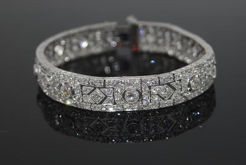 May include: A silver-toned metal bracelet featuring a repeating geometric pattern of small clear stones and larger circular stones set throughout the band. The bracelet has a hinged clasp mechanism and a reflective surface with multiple sparkling accents.