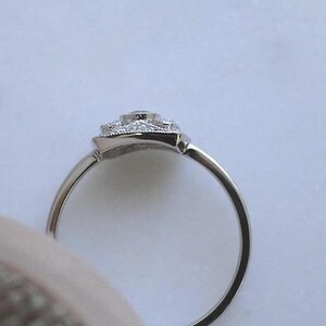 K&ouml;nnte beinhalten: Ein silberner Ring mit quadratischem, verziertem Besatz. Der Besatz ist mit kleinen, klaren Edelsteinen besetzt, die einen gr&ouml;&szlig;eren zentralen Stein umgeben. Der Ring ist schmal und poliert und reflektiert das Licht.