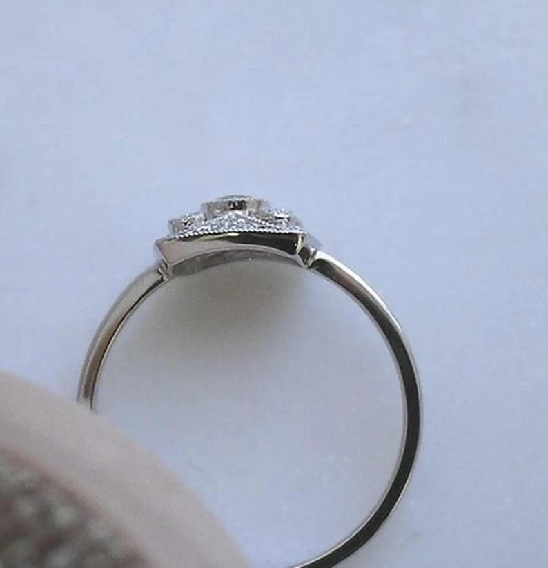 K&ouml;nnte beinhalten: Ein silberner Ring mit quadratischem, verziertem Besatz. Der Besatz ist mit kleinen, klaren Edelsteinen besetzt, die einen gr&ouml;&szlig;eren zentralen Stein umgeben. Der Ring ist schmal und poliert und reflektiert das Licht.