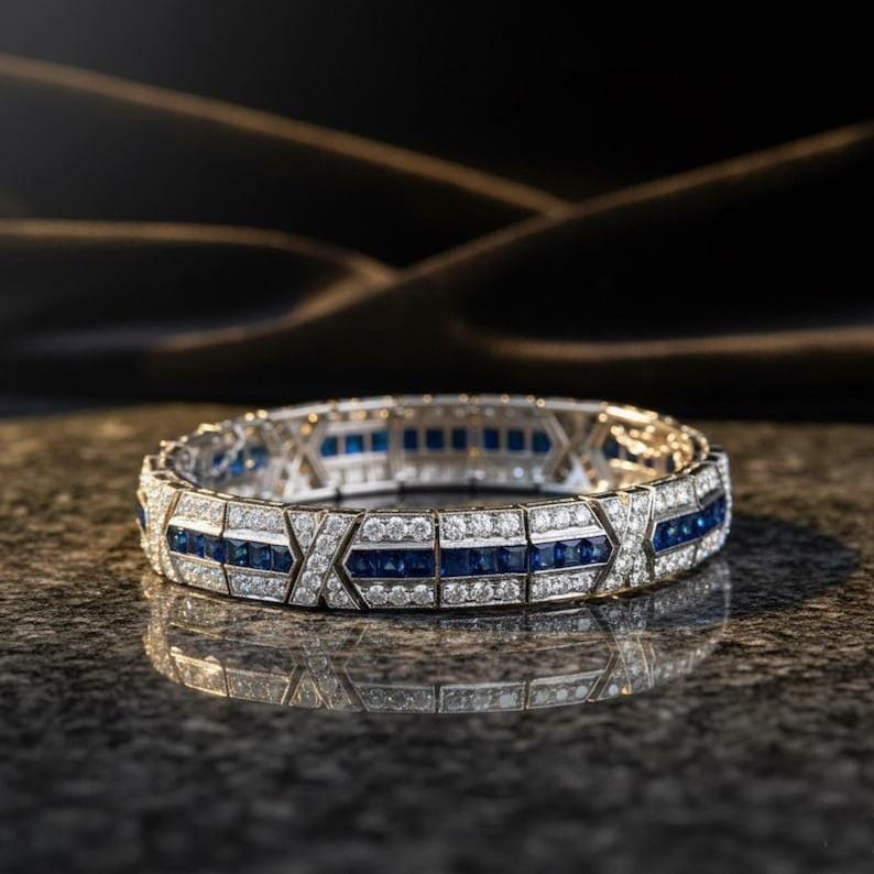 May include: A hinged bracelet featuring a central row of square-cut blue sapphires flanked by two rows of round brilliant-cut white diamonds. The metal band is segmented with X-shaped motifs and has a polished finish with a reflective surface.