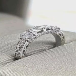 May include: A silver ring featuring three clear gemstones in a floral design. The band has detailed filigree patterns. The ring is displayed on a grey, textured surface. The ring's design is intricate.
