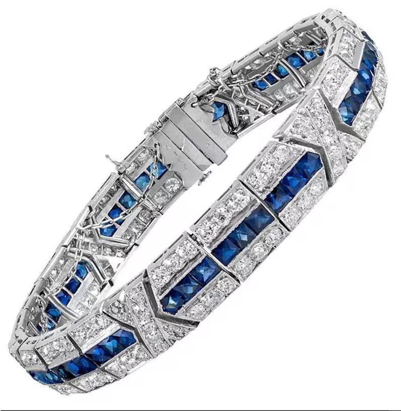 May include: A silver-toned metal bracelet featuring a central row of square-cut blue gemstones flanked by two rows of small, clear, round-cut stones. The design includes geometric metal segments and a secure clasp mechanism with a safety chain.