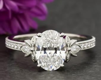 Anel de noivado com moissanita de corte oval em ouro branco 14k, aliança de casamento com flor-de-lis de inspiração vintage, aro cravejado com grãos e detalhe de pétala.