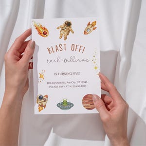 Peut inclure: Une invitation blanche sur le thème de l'espace, tenue par deux mains. La carte présente des illustrations d'une fusée, d'un astronaute et de planètes. Le texte indique "BLAST OFF!" et "Earl Williams A CINQ ANS !" avec les coordonnées.
