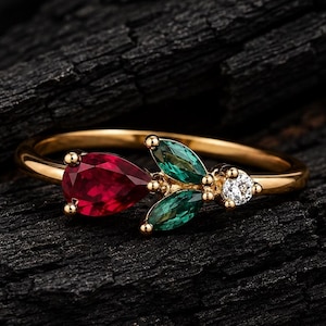 Peut inclure: Une bague en or ornée d'une pierre rouge en forme de poire, de deux pierres vertes taille marquise et d'une petite pierre claire ronde disposées en grappe. L'anneau est fin et en or poli, présentant une délicate composition florale.