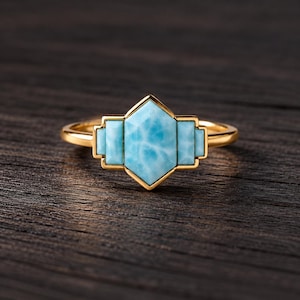 May include: A gold ring featuring a central hexagonal light blue stone flanked by two smaller rectangular light blue stones. The setting is crafted from polished gold metal, creating a geometric arrangement that highlights the marbled texture of the blue stones.