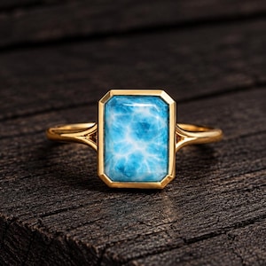 May include: A gold ring featuring a rectangular, light blue Larimar gemstone with a marbled, cloud-like pattern. The stone is set in a smooth, polished gold bezel, and the band features a split-shank design that connects to the setting.