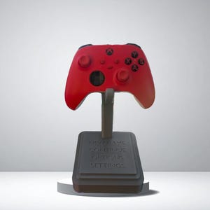 May include: A red Xbox controller is displayed on a black stand. The stand has a rectangular base with the text "NEW GAME CONTROLLER GAME SETTINGS". The controller is supported by a vertical post.