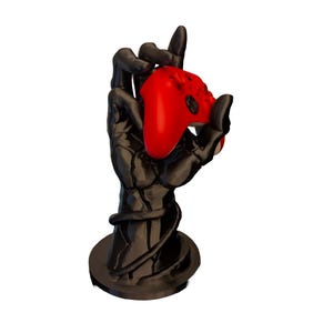 May include: A black, stylized hand-shaped stand holding a red video game controller. The stand has a circular base and a detailed, textured design. The controller is a vibrant red color.