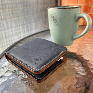Puede incluir: Una cartera de cuero negro con costuras y ribetes marrones, con la marca "DENRAVEN". La cartera está abierta, revelando el interior. Una taza verde claro con borde marrón se encuentra en el fondo.