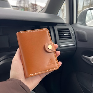 Könnte beinhalten: Ein braunes Lederportemonnaie mit weißen Nähten und einem goldenen Knopfverschluss. Das Portemonnaie trägt den Text "DEN RAVEN" und ein Vogellogo. Das Portemonnaie wird in einer Hand gehalten.