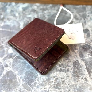 Peut inclure: Un portefeuille bifold en cuir marron foncé avec des coutures apparentes et un petit logo d'oiseau gaufré sur le devant. Une étiquette rectangulaire blanche avec un graphique d'empreinte de main noire est attachée par une fine ficelle.