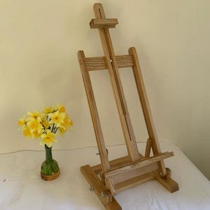 May include: A wooden artist's easel, made from light-coloured wood, is angled. The easel has adjustable features and a lower shelf. A small vase of yellow daffodils is nearby.