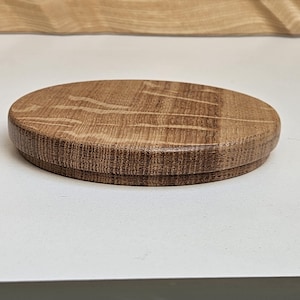 May include: A circular wooden lid with a natural wood grain. The lid is light brown and has a slightly raised edge. The wood grain is visible on the top and sides. The lid is likely for a container, and is made of oak.