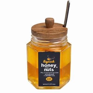 Peut inclure: Pot de miel hexagonal en verre avec un couvercle en bois et un petit bouton en bois. Une cuillère en métal est posée sur le couvercle. L'étiquette indique "honey & nuts". Le pot contient 230 grammes de miel.