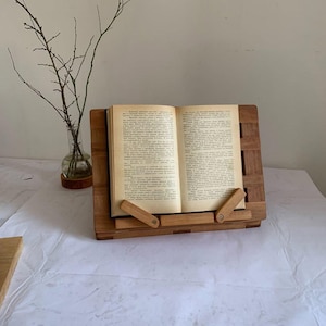 Könnte beinhalten: Ein hölzerner Buchständer mit einem offenen Buch. Der Ständer ist aus hellbraunem Holz gefertigt und hat ein Lattenmuster. Das Buch ist auf einer Textseite geöffnet. Eine Glasvase mit Zweigen befindet sich im Hintergrund.