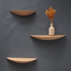 Könnte beinhalten: Drei schwebende Holzregale mit geschwungenem Design sind an einer grauen Wand befestigt. Eine kleine beige Vase mit getrockneten orangefarbenen Blumen steht auf dem mittleren Regal. Die Regale sind aus hellem Holz gefertigt.