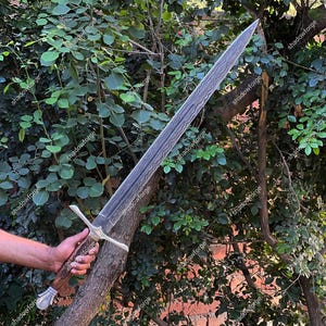 May include: A long, silver sword with a patterned blade and a cross-shaped hilt. The handle is a light brown color with a silver pommel. The sword is held against a backdrop of green foliage. The text "shadowforge" is visible on the image.