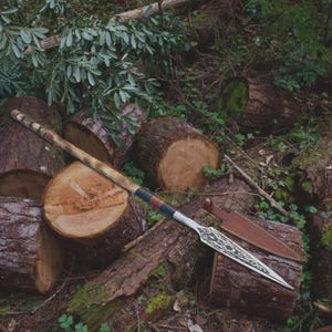 Puede incluir: Una lanza larga con una hoja de metal afilada y puntiaguda con intrincados grabados en tono plateado. El mango de madera está envuelto con detalles de cuero oscuro cerca de la base de la hoja. Una funda de cuero marrón a juego yace al lado.