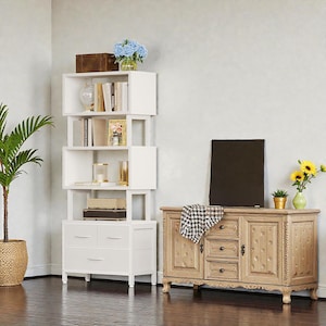 May include: A white, asymmetrical bookcase with open shelves and two drawers. The bookcase holds books, decorative items, and a framed picture. A wooden cabinet with drawers and doors is next to the bookcase. A potted plant and flowers are also in the room.