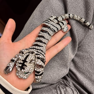 May include: A silver-toned articulated tiger brooch covered in clear rhinestones with black enamel stripes. The tiger features green rhinestone eyes and a flexible, segmented body design that allows for movement, showcasing a detailed feline head.