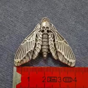 May include: A silver-toned metal moth brooch featuring a detailed skull design on the thorax and intricate wing patterns. The moth is positioned above a red measuring tape, showing a width of approximately 1.5 inches across the wingspan.