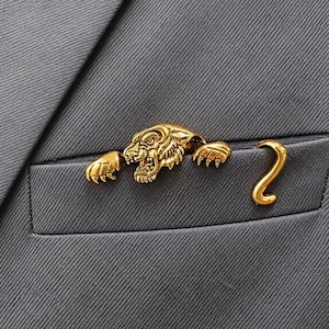 Peut inclure: Une broche en métal doré en forme de tigre, présentant une tête détaillée avec des crocs apparents et deux pattes avant étendues, accompagnée d'une pièce séparée en forme de queue courbée, fixée sur le bord d'une poche en tissu gris foncé.