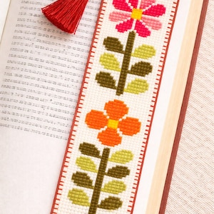 May include: A handmade bookmark featuring a cross-stitch floral design. The bookmark has a white background with two flowers in pink, orange, and yellow, with green leaves and brown stems. A red tassel hangs from the top.