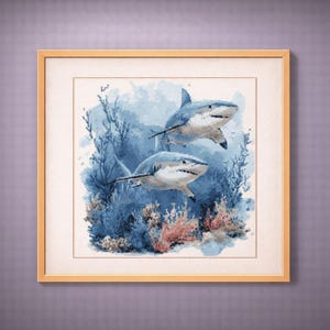 May include: Framed artwork featuring two white sharks swimming in a blue ocean scene. The sharks are depicted with detailed shading and open mouths, surrounded by blue coral and plants. The artwork is framed in a light brown wooden frame.