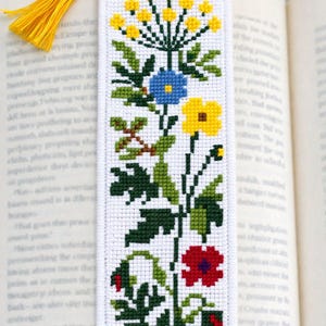 May include: White cross-stitch bookmark with a floral design. The bookmark features yellow, blue, red, and purple flowers with green leaves. A yellow tassel hangs from the top. The bookmark is placed in an open book.