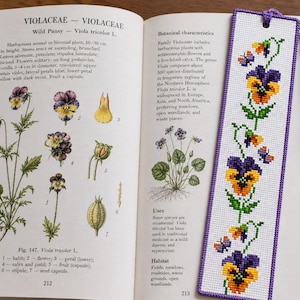May include: A rectangular cross-stitch bookmark featuring a vertical pattern of purple and yellow pansy flowers with green stems and leaves. The bookmark has a purple fabric border and a matching purple tassel attached to the top with a small hole.