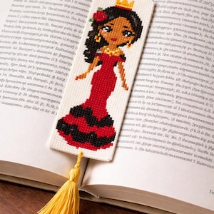 May include: Cross-stitch bookmark featuring a woman in a red dress with black accents, a gold crown, and a rose in her hair. The bookmark has a yellow tassel and is placed in an open book.