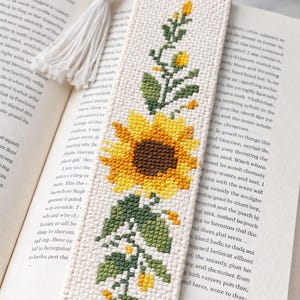 May include: Cream-coloured embroidered bookmark featuring a sunflower design. The bookmark has a yellow and brown sunflower, green leaves, and a white tassel. It is in an open book.