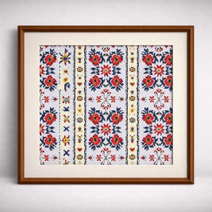 May include: Framed artwork featuring a detailed cross-stitch pattern. The design showcases a repeating floral motif in red, blue, and yellow on a white background. The artwork is encased in a brown wooden frame with a white mat.
