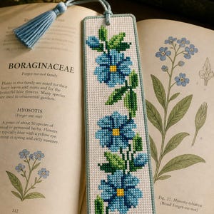 May include: A handmade bookmark featuring a cross-stitched design of blue forget-me-not flowers with green leaves and yellow centers. The bookmark has a light blue border and a matching tassel. It rests on an open book with botanical illustrations.