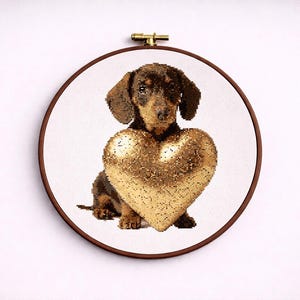 May include: A cross-stitch embroidery of a dachshund dog holding a gold heart. The dog is brown and black, and the heart is a shimmering gold. The embroidery is set in a round wooden hoop.
