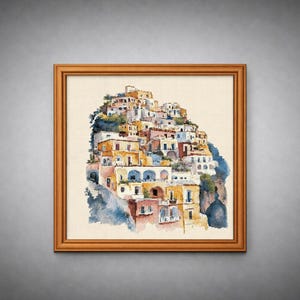 Puede incluir: Pintura a la acuarela de un pueblo costero italiano, con edificios coloridos en una ladera. La obra de arte está enmarcada en un marco de madera sobre un fondo gris. Los edificios están pintados en tonos de amarillo, rosa y azul.