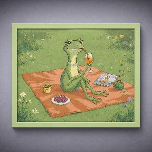 May include: Framed artwork featuring a green frog enjoying a picnic. The frog is wearing a necklace and holding a drink. A red blanket, food, and a book are also visible. The artwork is in a green frame.