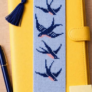 Op de afbeelding: Blauw kruissteek bladwijzer met vier blauwe en roze zwaluwen. De bladwijzer heeft een blauwe kwast en ligt op een geel notitieboek. Een blauwe pen en plakbriefjes zijn ook zichtbaar.