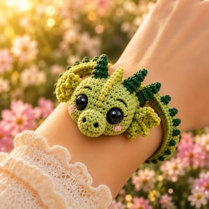 May include: A crochet bracelet featuring a green dragon head with dark green horns and spikes, large black glossy eyes, and small pink blush marks on the cheeks. The band is made of green yarn with a repeating dark green spike pattern along the edge.