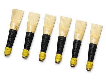 May include: Six bassoon reeds arranged in a row, each featuring a light beige cane tip, a black thread-wrapped middle section, and a bright yellow thread-wrapped base with a small metal tube opening at the bottom end of each individual reed.