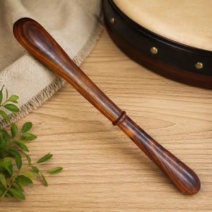 May include: A wooden bodhrán beater with a rounded end and a ring detail. The beater is a rich brown colour with visible wood grain. The background includes a bodhrán drum and a wooden surface.