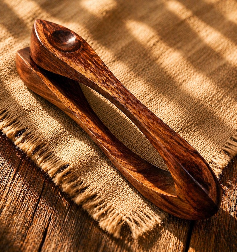 Op de afbeelding: Een paar houten serveertangen, gemaakt van donkerbruin hout met zichtbare nerfpatronen. De tangen hebben een afgeronde handgreep en een lepelvormig uiteinde. Ze liggen op een textiel, beige stof, wat een rustieke of natuurlijke uitstraling suggereert. (ongeveer 20 cm)