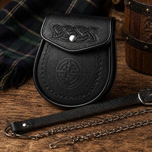 May include: A black leather sporran with Celtic knot detailing on the flap and a circular emblem on the body. It features a leather strap and chain. The sporran is presented on a wooden surface, with tartan fabric visible.