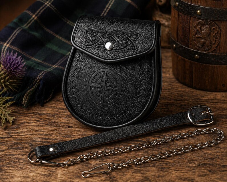 May include: A black leather sporran with Celtic knot detailing on the flap and a circular emblem on the body. It features a leather strap and chain. The sporran is presented on a wooden surface, with tartan fabric visible.